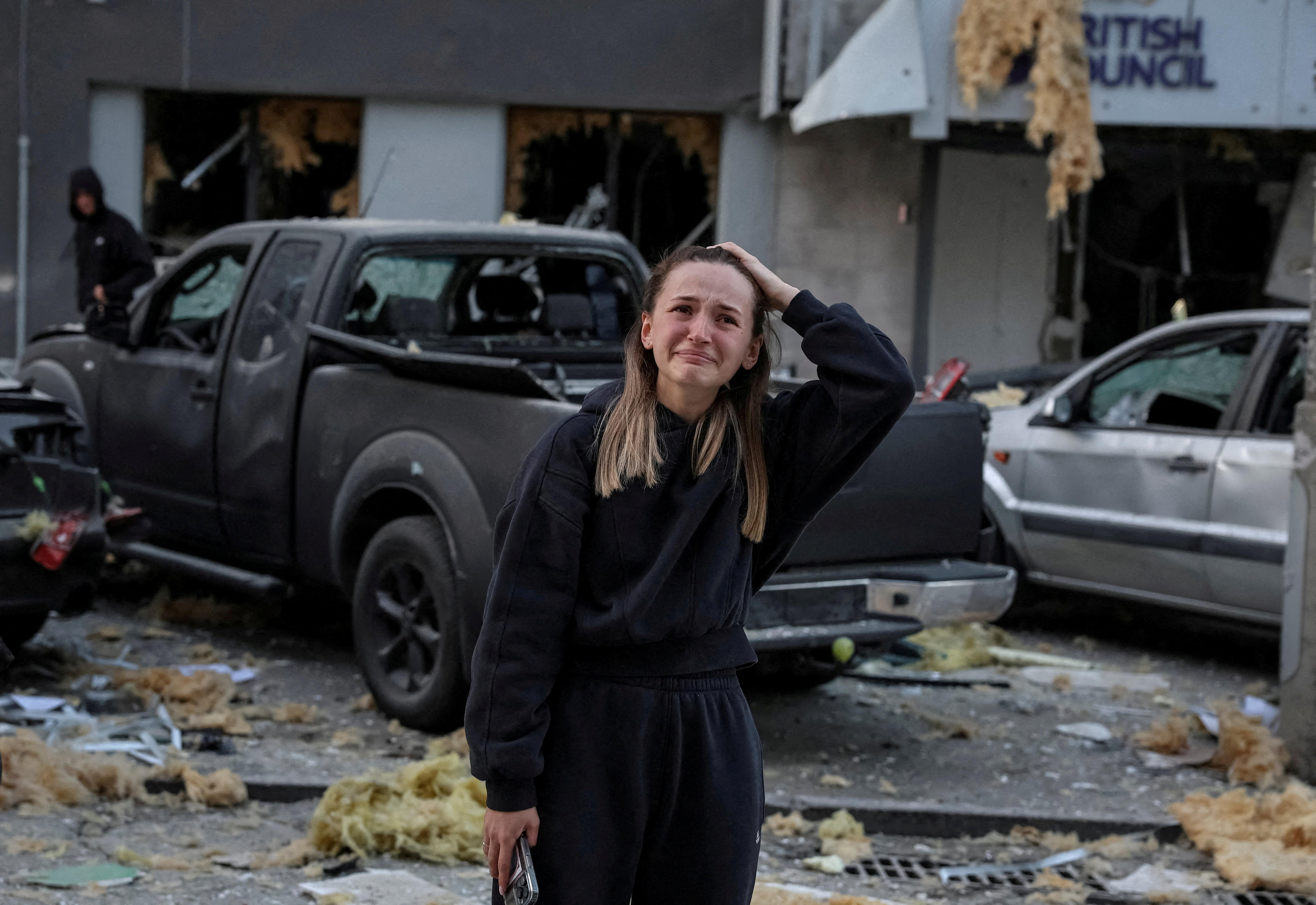 A crying woman stands in front of damaged cars and building.