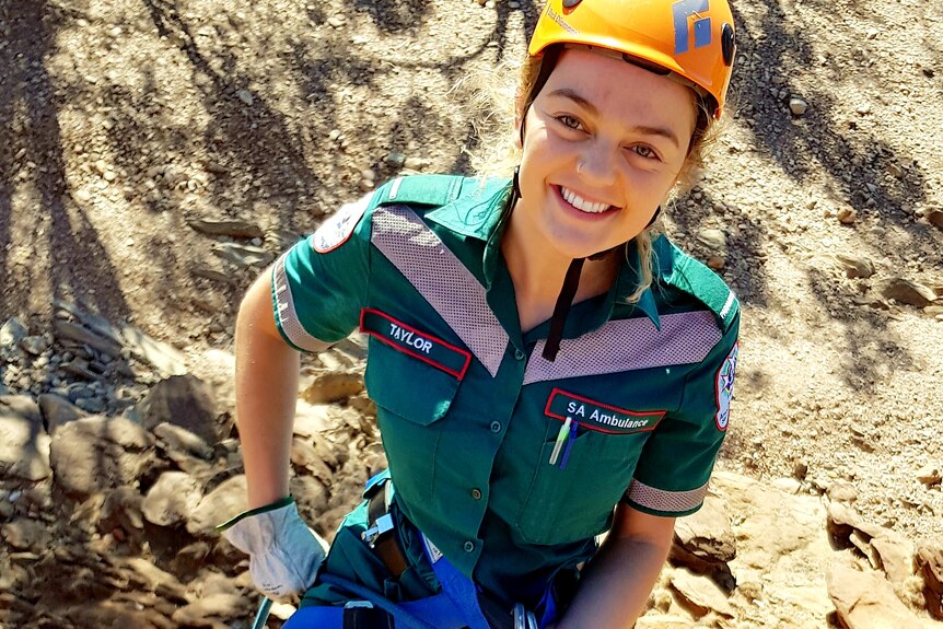 A woman wearing a green ambulance uniform and a helmet climbing up a rock cliff. 