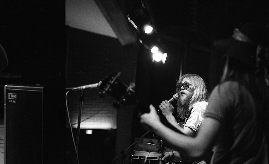 Black and white 70s photo of a long-haired, bearded man in round sunglasses singing, bass player out of focus in the foreground.