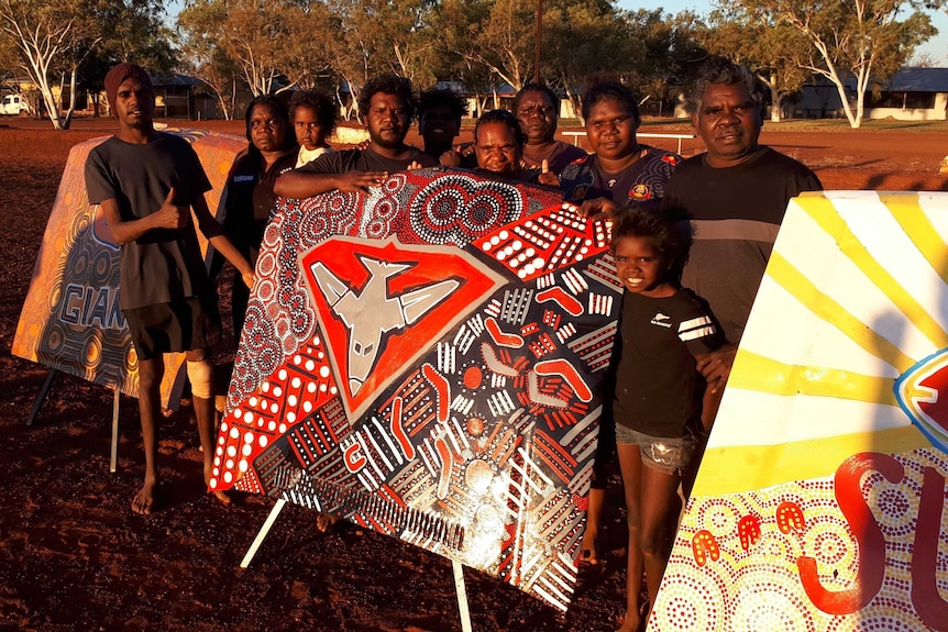 A group of Indigenous people wearing dark clothing stand around a brightly-painted car bonnet.