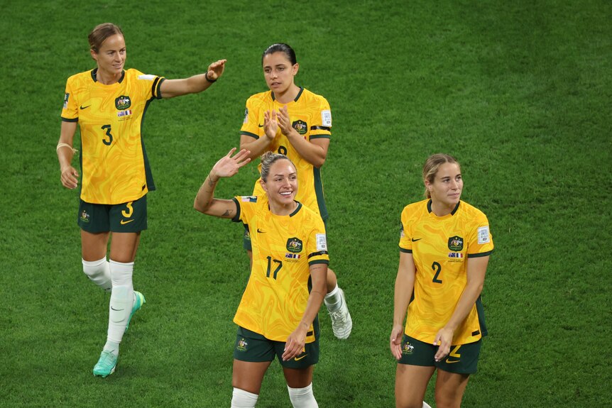 Women soccer players in gold shirts wave to crowd.