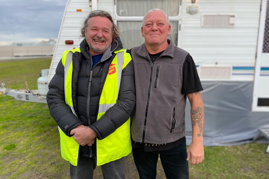 Two men stand outside a caravan smiling