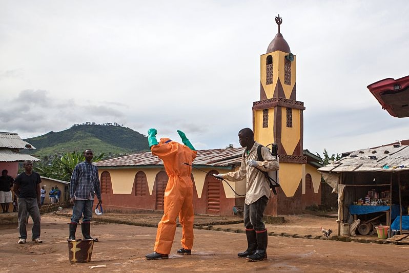 A man in an orange protective suit is sprayed with chlorine by a man in ordinary clothes.