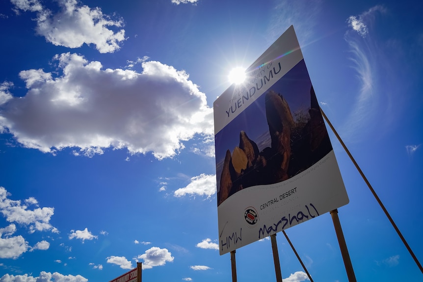 A welcome sign to the remote community of Yuendumu, in front of blue sky, sun and a few clouds.