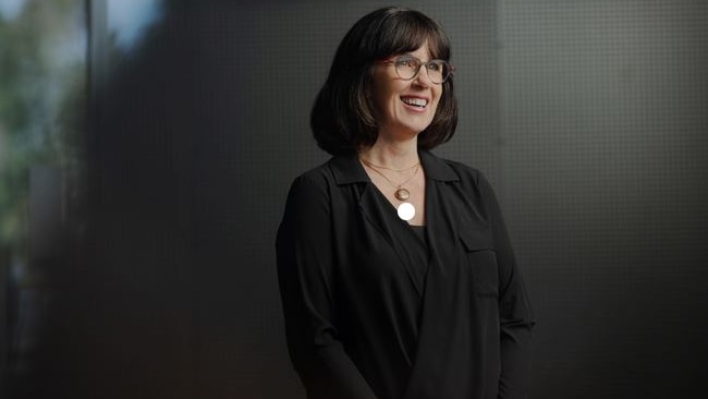 Woman wearing dark clothes, with dark hair and glasses looks into the distance and smiles.