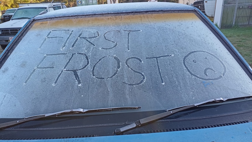 A frosty car windscreen with "first frost" and a sad face drawn into the ice.