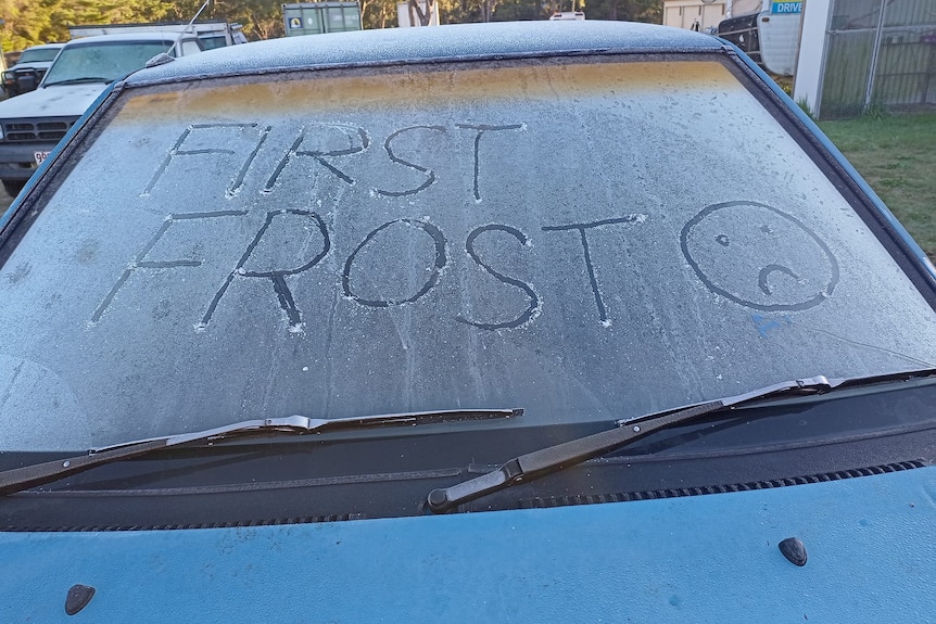 A frosty car windscreen with "first frost" and a sad face drawn into the ice.