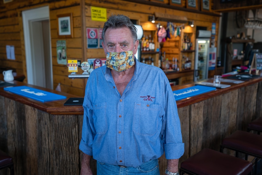 A man wearing a covid face mask looking at the camera in front of a bar.