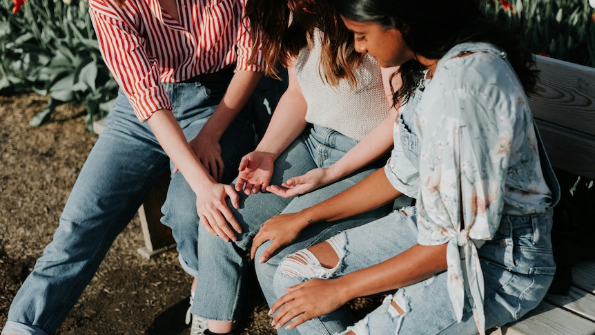 Three women sitting on a grey bench wearing blue jeans. 
