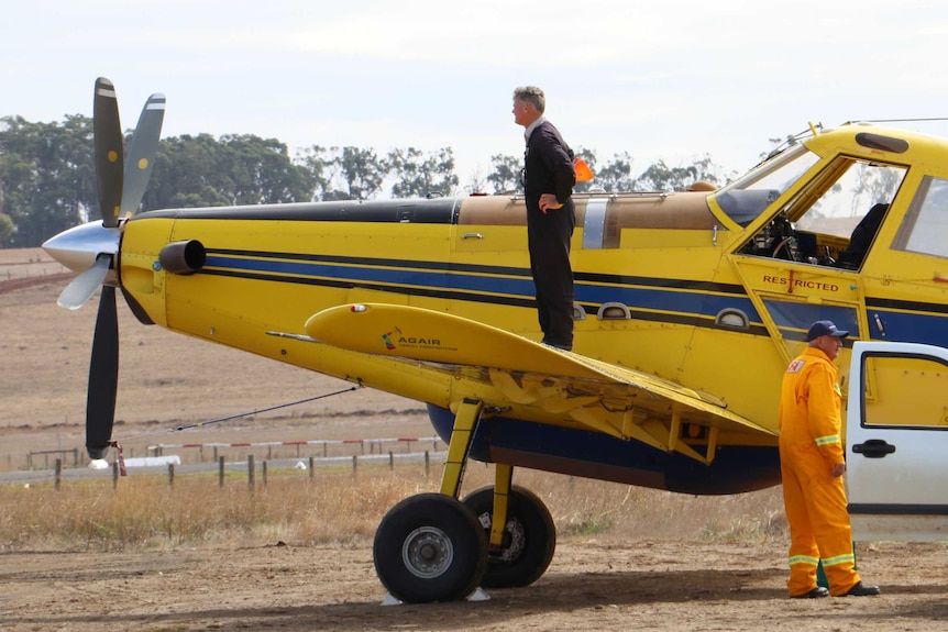 A man stands on the wing of a plane at the Cobden airport, looking off to the distance. A CFA volunteer stands nearby.