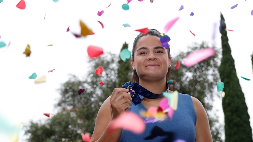 Madison Prespakis holds up her medal with trees in the background and confetti falling.