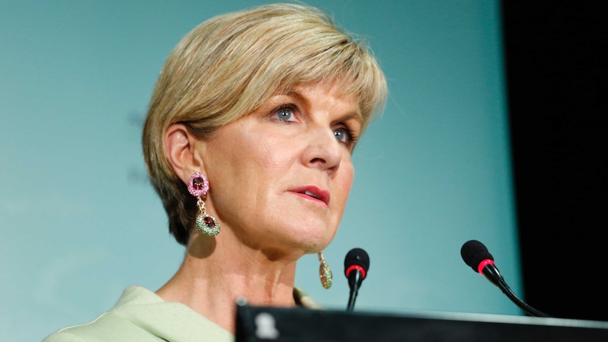 Julie Bishop speaks into two microphones at a lectern. She is wearing a beige coloured suit and jewelled earrings.