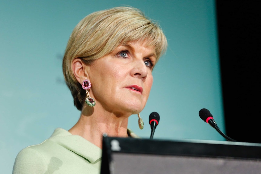 Julie Bishop speaks into two microphones at a lectern. She is wearing a beige coloured suit and jewelled earrings.