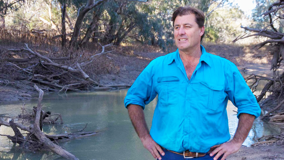 A man stands with his hands on his hips, before a river that looks low on water, surrounded by sticks.