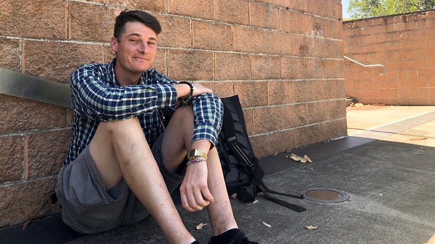 A thin young man grins as he sits on the ground leaning against a wall signposted with TAFE NSW Illawarra institute.