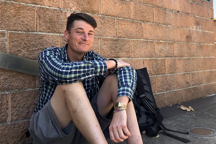 A thin young man grins as he sits on the ground leaning against a wall signposted with TAFE NSW Illawarra institute.