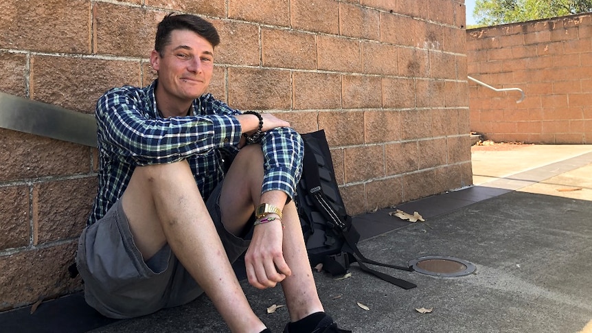 A thin young man grins as he sits on the ground leaning against a wall signposted with TAFE NSW Illawarra institute.