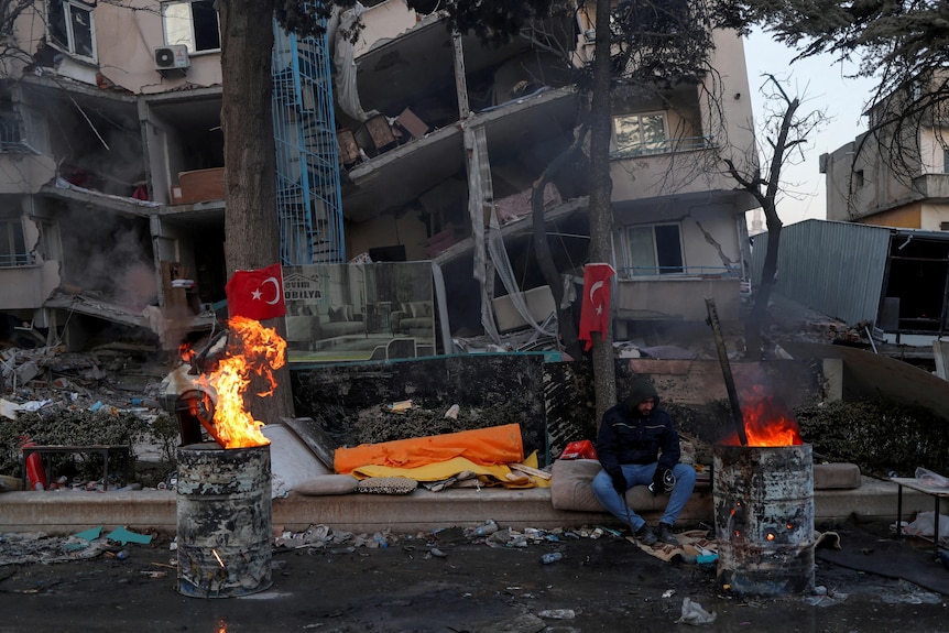 A man sits outside a building in front of a fire. 