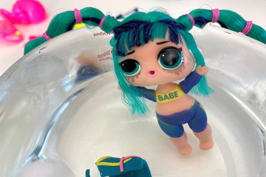 A small plastic doll sitting in bowl of water with bike pants and a top with "babe" written across it.