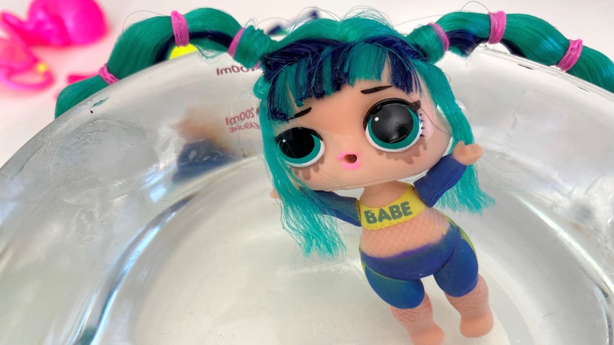 A small plastic doll sitting in bowl of water with bike pants and a top with "babe" written across it.