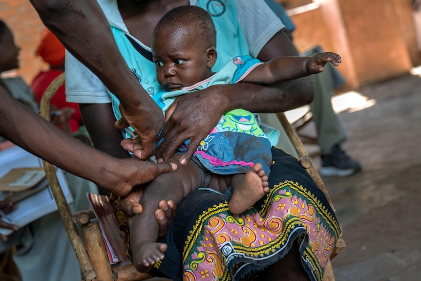A young African baby receives an injection in his thigh as he is held by his mother who is seated on a chair.