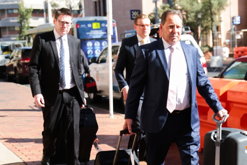 Defence lawyer David Edwardson walks ahead of two other men in suits carrying two pieces of luggage.