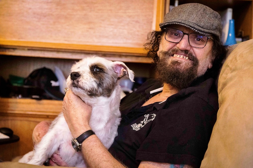 A man wearing a cap and a black shirt, smiling with a dog sitting on his lap.