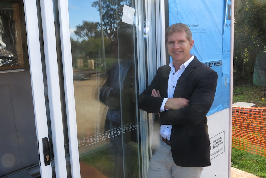 A man with a suit, with his arms crossed, standing in front of a sliding class door on a tiny house