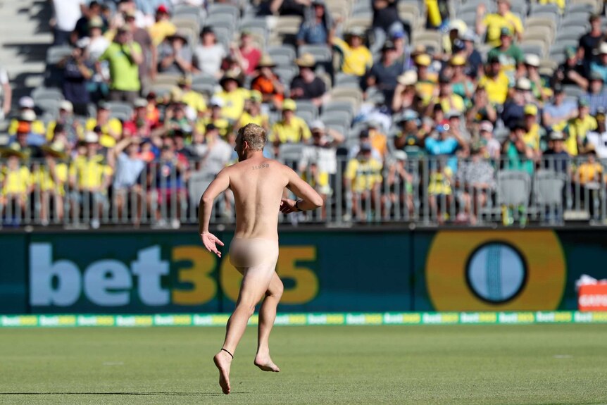 A streaker wearing no clothes runs on the playing surface at Perth Stadium in front of a crowd in the stands.