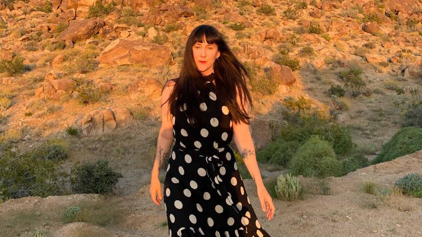 A woman in a black dress in white spots with sun on desert rocks in the background. 