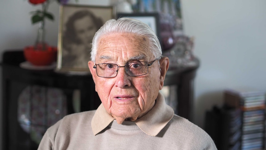 An elderly war veteran looks at the camera, with framed pictures and a flower in the background.