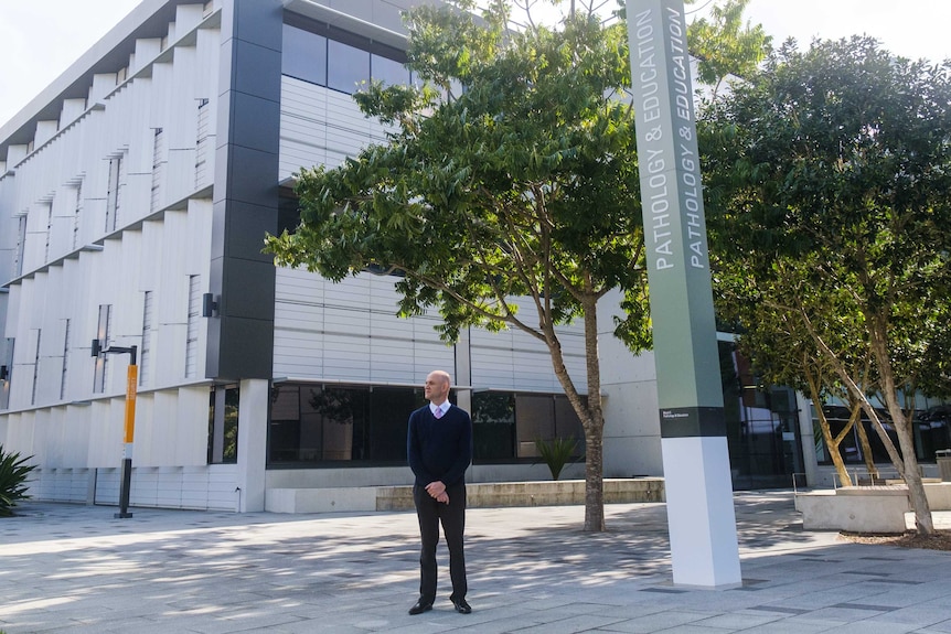 A man stands in a shady spot on a hospital campus with a sign behind him reading "Pathology and Education".