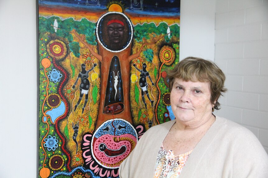 Woman in a cream cardigan standing in front of an indigenous artwork with greens, blues browns and pink