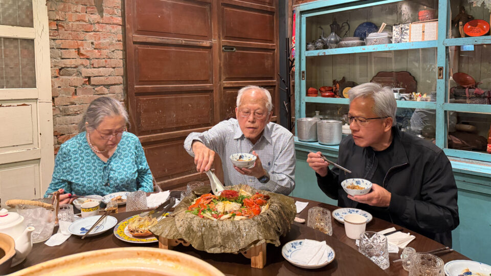 Jensen Huang in a black jacket eats from a bowl with chopsticks at a table with an elderly man and woman.