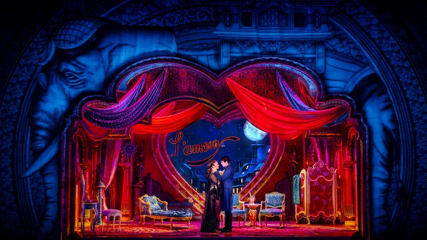 On a stage, a man and woman embrace in front of an elaborate Bohemian Parisian set