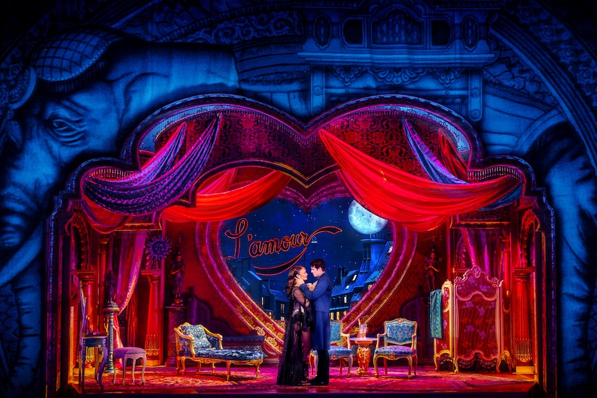 On a stage, a man and woman embrace in front of an elaborate Bohemian Parisian set