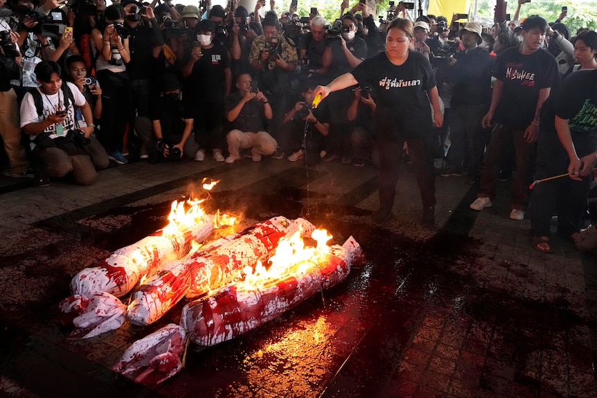 A woman surrounded by a crowd of people sets alight effigies splattered with red paint. 