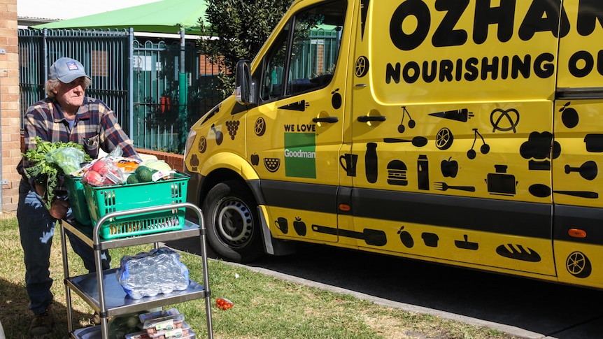 A man pushes a trolley filled with food next to a yellow OzHarvest truck parked on the side of the street.
