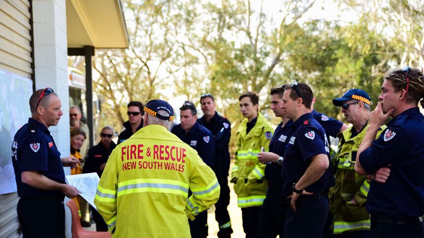 Firefighters stand around listening to a briefing, the closest to camera is wearing a bright florescent jacket.