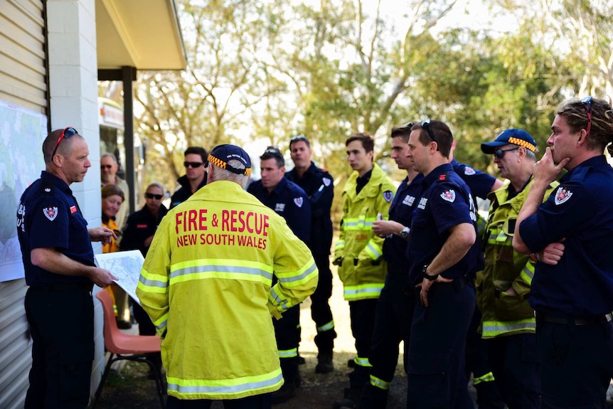 Firefighters stand around listening to a briefing, the closest to camera is wearing a bright florescent jacket.