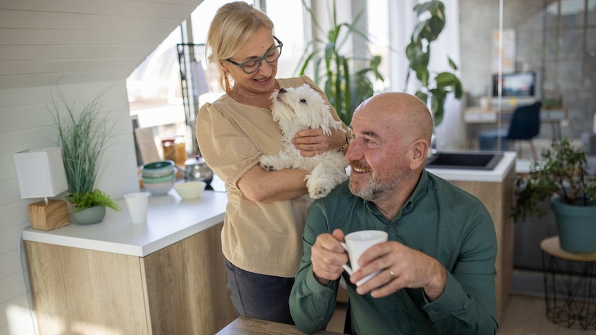 A mature couple smile in their small kitchen/ dining space with a cuddly maltese