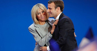 Emmanuel Macron and his wife Brigitte, both dressed in suit jackets, embracing and smiling before a bright blue background.