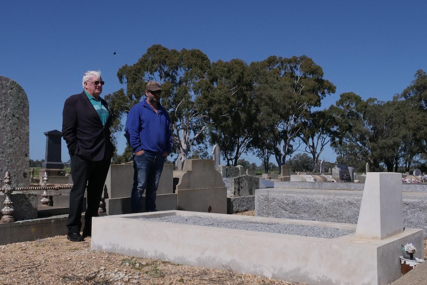 Two men stand under a blue sky in a graveyard looking over a grave