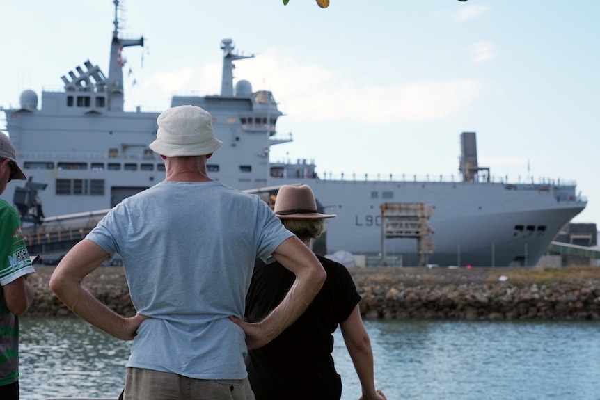 A woman and a man with his hands on his hips watch a large naval ship 