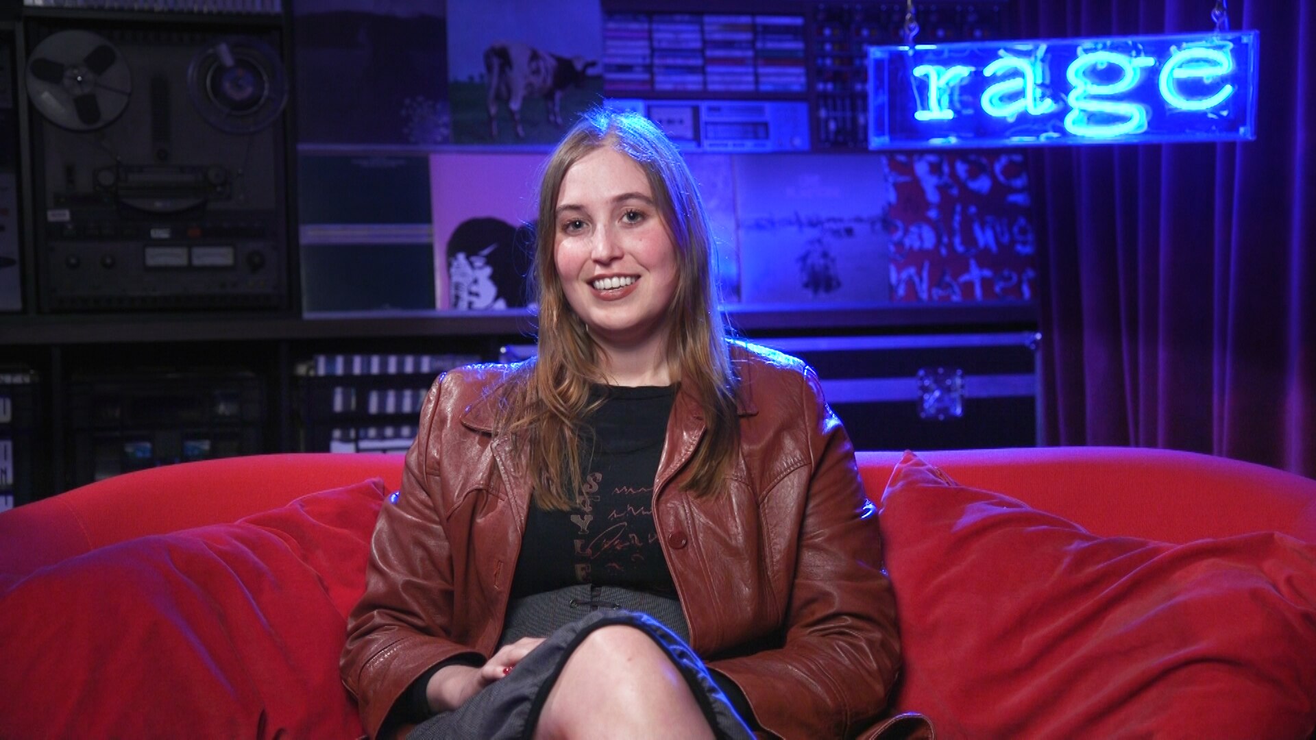 Hatchie sits smiling on a red couch in front of a blue neon sign that reads rage