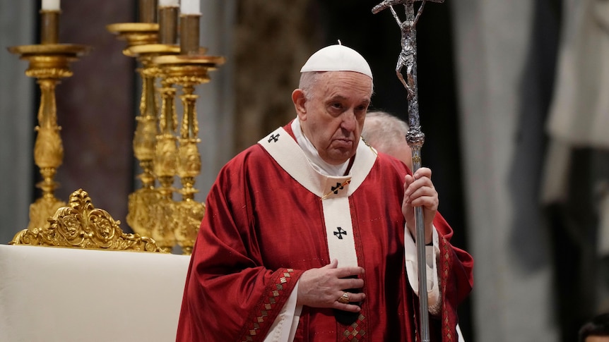 The Pope wears a white skullcap and red robes as he holds a staff featuring a carving of Jesus being crucified.