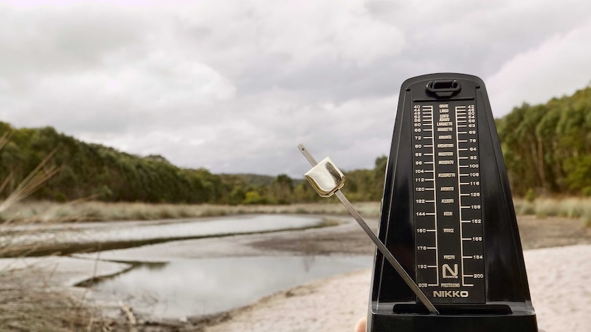A hand holds a metronome in front of a river landscape