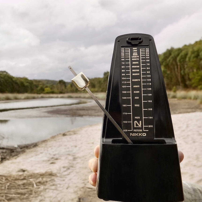 A hand holds a metronome in front of a river landscape