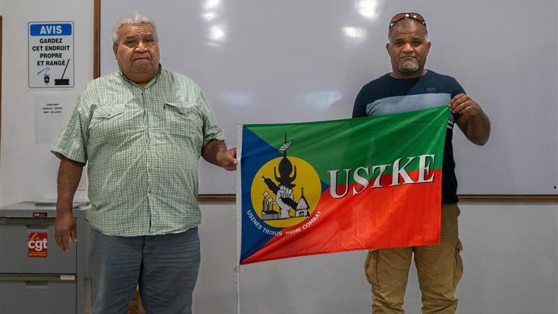 Two men hold up colourful flag in front of white board.