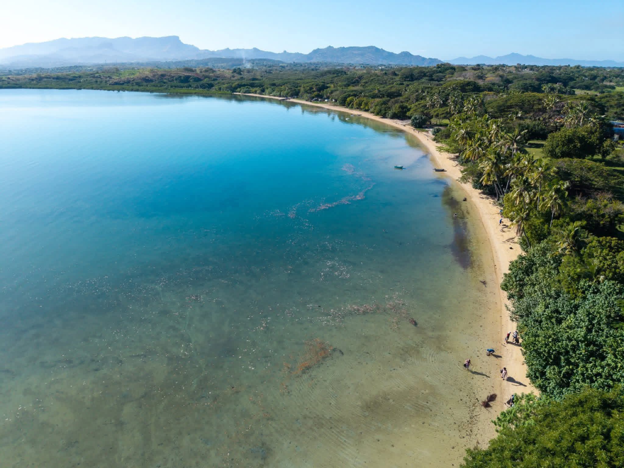 Drone photos show a vast bay with crystal blue water lapping against a white sandy beach with palm trees lining the shoreline.